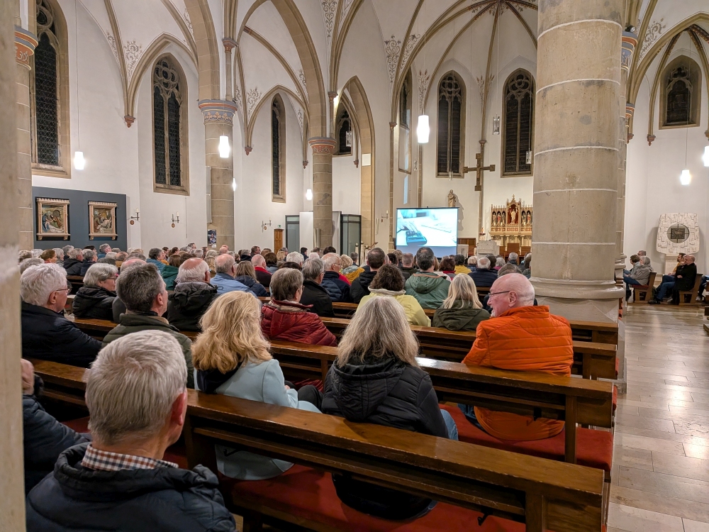 Bild vieler Menschen in einer Kirche. Von hinten aufgenommen. Vorn ist eine Leinwand mit einer Powerpointpräsentation zu sehen. 